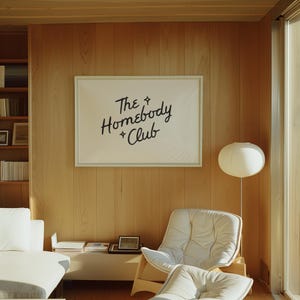 May include: A light-filled interior features a framed artwork with the text "The Homebody Club" in black script. The room includes a white sofa, a modern chair, and a floor lamp with a round shade. The walls are paneled with light-colored wood.