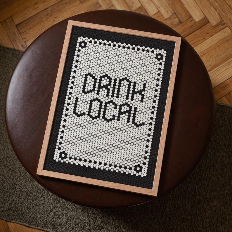 May include: A framed art print with the words "DRINK LOCAL" in black letters. The artwork features a white and black mosaic tile design with a wooden frame. The print is displayed on a brown, circular ottoman.