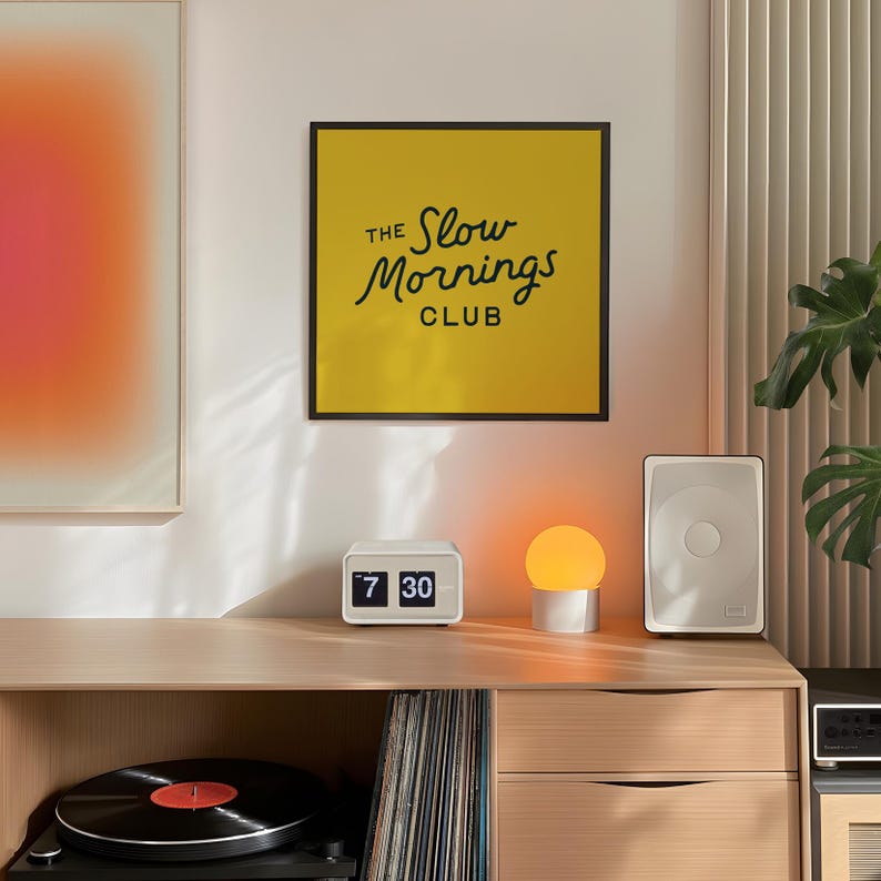 以下が含まれることがあります： 「The Slow Mornings Club」のテキストが入った、額装された黄色のプリントが特徴的なモダンなリビングルーム。白いフリップクロックは7:30を表示し、小さなオレンジ色のグローブランプが、レコードプレーヤーとスピーカーを備えた木製のキャビネットの上に置かれています。