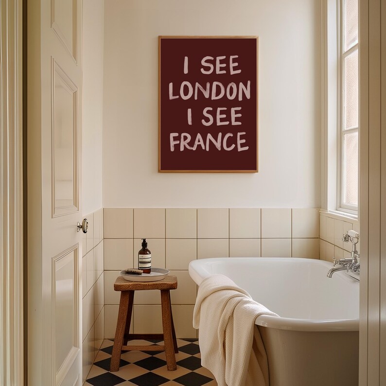 May include: A bathroom scene featuring a framed print with the text "I SEE LONDON I SEE FRANCE" in white on a maroon background. The room includes a white bathtub, a wooden stool, and a bottle of soap. The floor has a black and white checkered pattern.