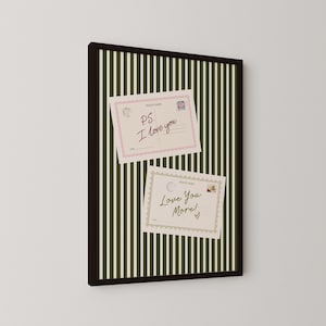 May include: A framed wall art piece featuring a dark olive green and white vertical striped background. Two rectangular postcards are displayed on the stripes. The top postcard has a pink border and the text P.S. I love you. The bottom postcard has a scalloped border and the text Love You More! with a small heart.