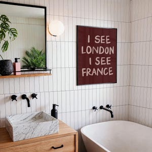 May include: A modern bathroom with white tile walls and a wooden vanity. A framed print on the wall reads "I SEE LONDON I SEE FRANCE". The bathroom features a marble sink, black faucets, and a white bathtub.