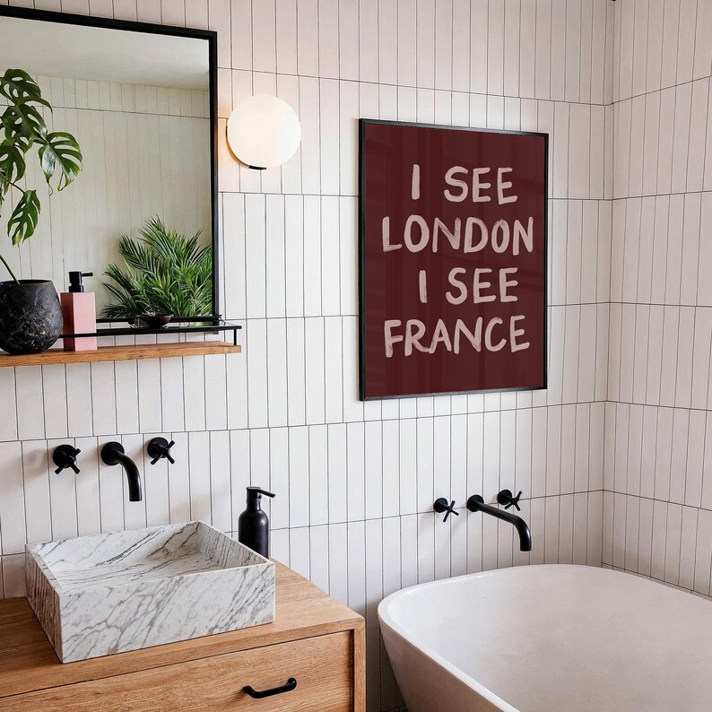 May include: A modern bathroom with white tile walls and a wooden vanity. A framed print on the wall reads "I SEE LONDON I SEE FRANCE". The bathroom features a marble sink, black faucets, and a white bathtub.