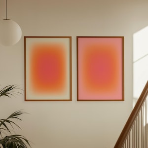 Pode incluir: Duas impressões de arte abstrata emolduradas com um gradiente de tons laranja e rosa. A obra de arte é exibida em uma parede branca, com uma escada e uma luminária suspensa visíveis. Uma planta em vaso está em primeiro plano.