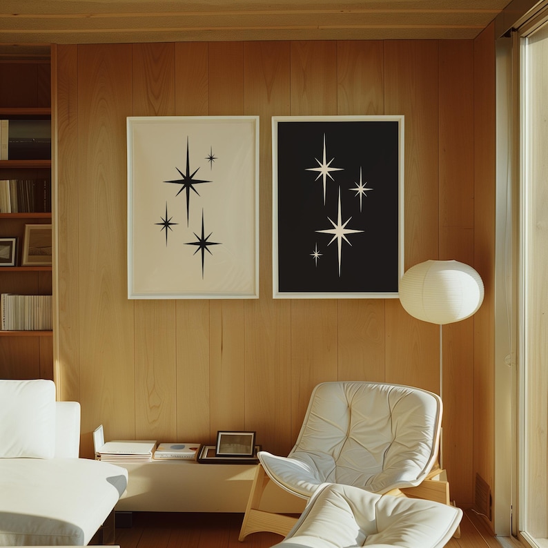 May include: Two framed starburst art prints. One print has a cream background with black starbursts, and the other has a black background with cream starbursts. The prints are displayed on a wood-paneled wall above a white chair and a white sofa.