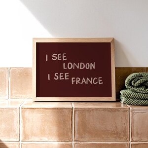 May include: A rectangular wall art print in a light wood frame featuring a dark maroon background with the text I SEE LONDON I SEE FRANCE written in a light pink, hand-drawn style font. The print is displayed in a horizontal orientation.