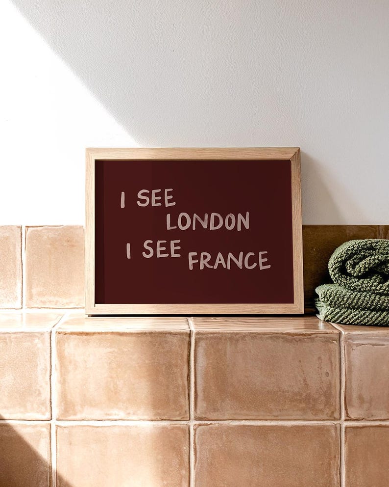 May include: A rectangular wall art print in a light wood frame featuring a dark maroon background with the text I SEE LONDON I SEE FRANCE written in a light pink, hand-drawn style font. The print is displayed in a horizontal orientation.