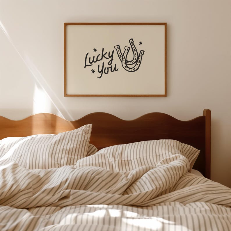 Może przedstawiać: Oprawiony wydruk z napisem "Lucky You" i ilustracją podkowy w kolorze czarnym. Grafika wisi nad ł&oacute;żkiem z pościelą w paski i drewnianym wezgłowiem. Pok&oacute;j ma neutralną paletę kolor&oacute;w.
