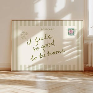May include: A framed postcard print with the words "it feels so good to be home" in a handwritten style. The postcard has a vintage aesthetic with a stamp and a Paris postmark. The background is a light beige with vertical stripes.