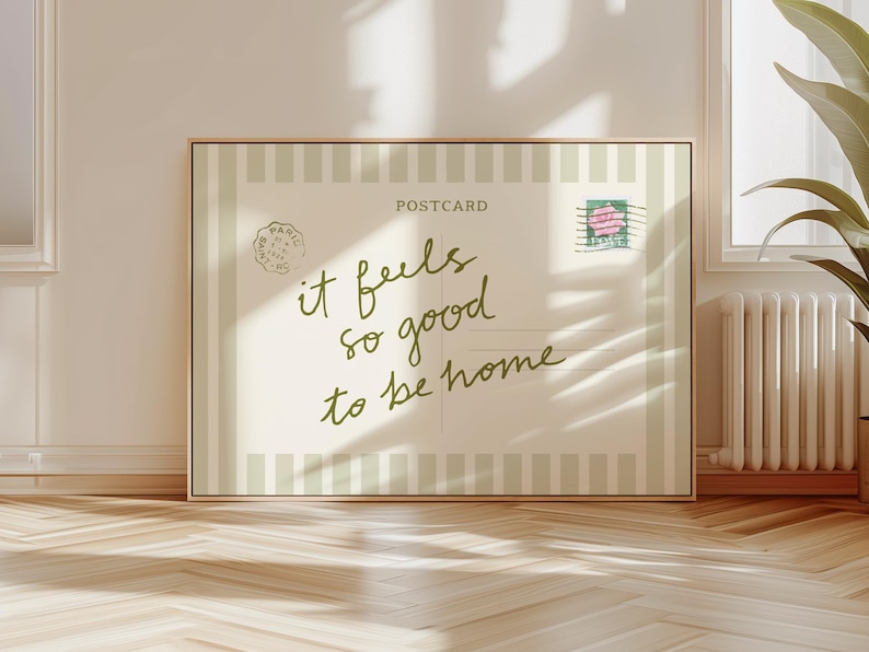 May include: A framed postcard print with the words "it feels so good to be home" in a handwritten style. The postcard has a vintage aesthetic with a stamp and a Paris postmark. The background is a light beige with vertical stripes.