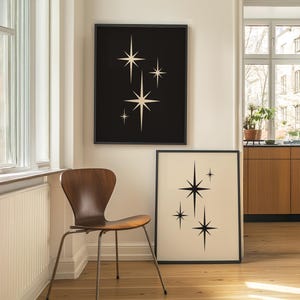 May include: Two framed art prints featuring starburst designs. One print has a black background with cream-colored stars, and the other has a cream background with black stars. The prints are displayed in a room with a wooden chair.