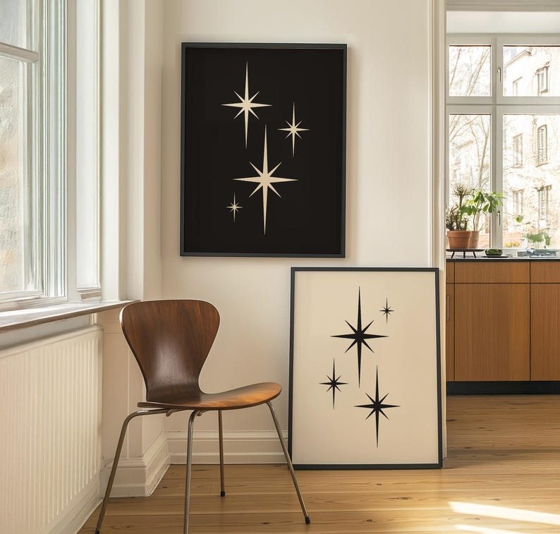 May include: Two framed art prints featuring starburst designs. One print has a black background with cream-colored stars, and the other has a cream background with black stars. The prints are displayed in a room with a wooden chair.