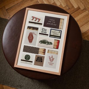 May include: A framed art print featuring various matchbox designs. The designs include text like "777", "The End", "Lucky You", and "This is a High Class Joint". The color palette includes red, white, black, and brown.