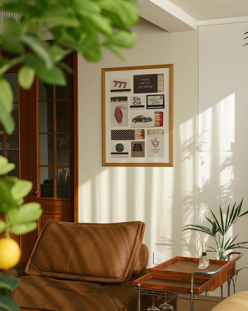 May include: A framed artwork featuring various vintage-style images, including a hotel key tag, a classic car, and matchboxes with text. The artwork is displayed on a light-colored wall, with a brown leather sofa and a wooden serving cart in the foreground.