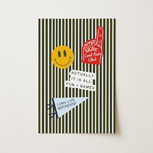 May include: A decorative card with a green and white striped background. The card features a yellow smiley face, a red foam finger with "Cool Kids Club" text, a white sticker with "ACTUALLY, IT IS ALL FUN + GAMES" text, and a blue pennant with "LONG LIVE BOYHOOD" text.