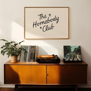 May include: A mid-century modern wooden cabinet with a record player, vinyl records, and a framed print that reads "The Homebody + Club." A potted plant sits on the cabinet. The album cover of The Beatles' Abbey Road is visible.