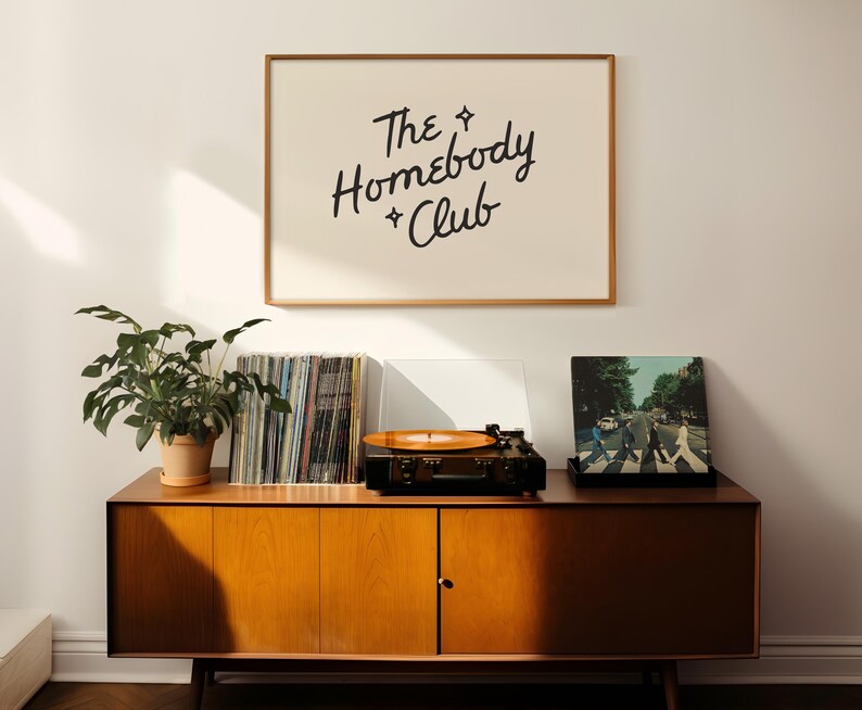 May include: A mid-century modern wooden cabinet with a record player, vinyl records, and a framed print that reads "The Homebody + Club." A potted plant sits on the cabinet. The album cover of The Beatles' Abbey Road is visible.
