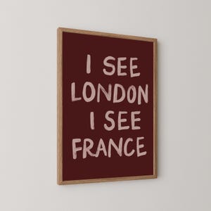 May include: A framed art print with the text "I SEE LONDON I SEE FRANCE" in white on a burgundy background. The frame is made of light brown wood. The artwork is displayed against a white wall.