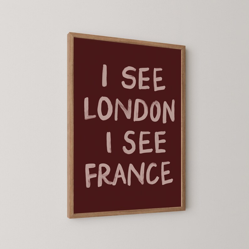 May include: A framed art print with the text "I SEE LONDON I SEE FRANCE" in white on a burgundy background. The frame is made of light brown wood. The artwork is displayed against a white wall.