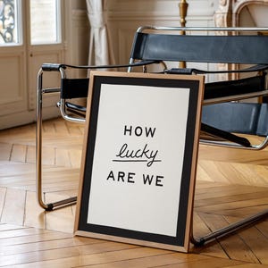 Puede incluir: Un cuadro enmarcado con el texto "HOW lucky ARE WE" en negro sobre fondo blanco. El marco es de color madera natural con un borde negro. El cuadro est&aacute; apoyado contra una silla sobre un suelo de parquet.