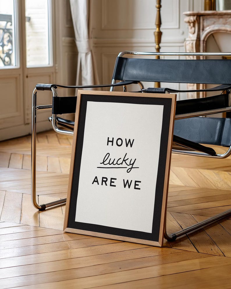 Puede incluir: Un cuadro enmarcado con el texto "HOW lucky ARE WE" en negro sobre fondo blanco. El marco es de color madera natural con un borde negro. El cuadro est&aacute; apoyado contra una silla sobre un suelo de parquet.