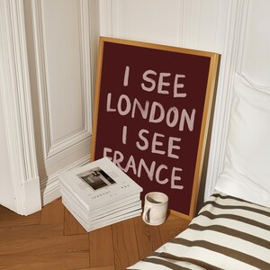 May include: A framed burgundy sign with the text "I SEE LONDON I SEE FRANCE" in white. A stack of white journals and a small ceramic mug sit on the wooden floor. The scene is next to a white wall and a bed with a striped comforter.