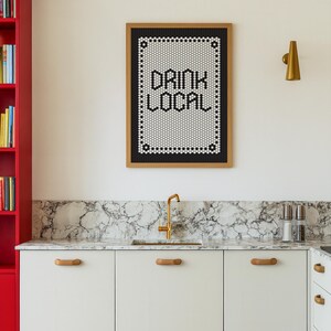 May include: A kitchen scene featuring a framed sign that reads "DRINK LOCAL" in black letters. The sign is made of small white and black tiles. Below, white cabinets with wooden handles and a marble countertop are visible.