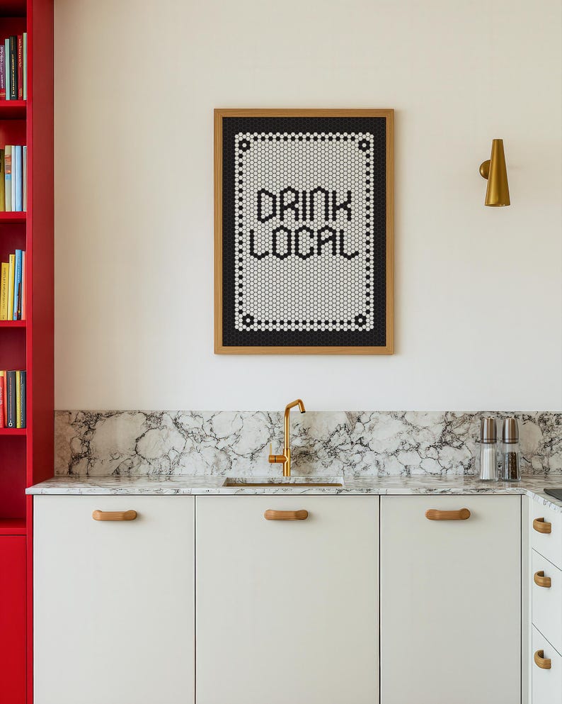 May include: A kitchen scene featuring a framed sign that reads "DRINK LOCAL" in black letters. The sign is made of small white and black tiles. Below, white cabinets with wooden handles and a marble countertop are visible.