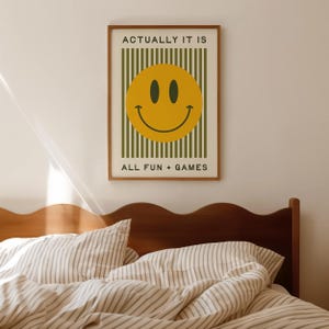 May include: A framed poster with a yellow smiley face design. The poster has the text "ACTUALLY IT IS" at the top and "ALL FUN + GAMES" at the bottom. The background is white with green vertical stripes.