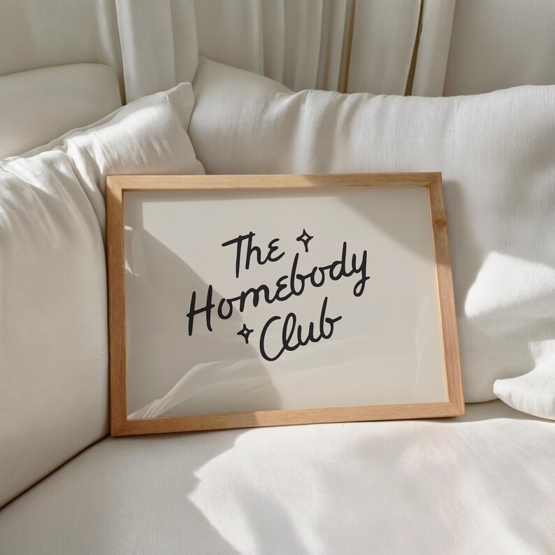 May include: A rectangular wooden framed print with the text "The Homebody Club" in black script. The print has a cream-colored background and is resting on a white surface.
