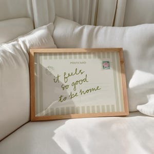 May include: A framed postcard with the text "it feels so good to be home" in a handwritten style. The postcard has a light beige background with vertical stripes and a postage stamp. The frame is made of light-colored wood.