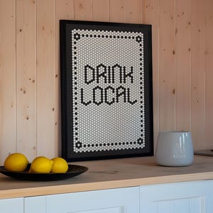 May include: A framed sign with the words "DRINK LOCAL" in black block letters. The sign has a black frame and a white background with a black and white mosaic tile design. Three yellow lemons sit in a black bowl.