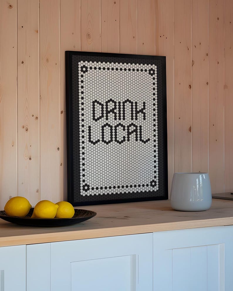 May include: A framed sign with the words "DRINK LOCAL" in black block letters. The sign has a black frame and a white background with a black and white mosaic tile design. Three yellow lemons sit in a black bowl.
