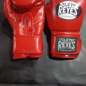 Peut inclure: Une paire de gants de boxe rouge vif. Les gants ont des coutures blanches et la marque "CLETD REYES" est affichée sur la sangle de poignet et le dessus du gant. Les gants sont fabriqués au Mexique.