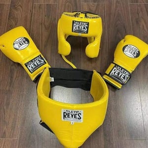 May include: A complete boxing set in yellow, including gloves, headgear, and a protective cup. Each piece features the "CLETD REYES" logo in black and white. The set is displayed on a wooden surface.