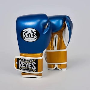 Peut inclure: Paire de gants de boxe bleus et dorés. Les gants ont un extérieur bleu avec des détails dorés sur le poignet et la paume. La marque "CLET0 REYES" est bien visible. Ces gants sont conçus pour un usage professionnel.