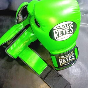 Puede incluir: Guantes de boxeo verde brillante con detalles en negro. Los guantes presentan el logotipo "CLET0 REYES" en blanco y negro. Fabricados en cuero, incluyen una correa de muñeca para un ajuste seguro.