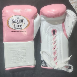 Puede incluir: Par de guantes de boxeo rosas y blancos. Los guantes tienen acolchado rosa en los nudillos y la muñeca, con cuero blanco en la palma y el puño. El texto "NO BOXING NO LIFE" está impreso en los guantes. Hecho en México.