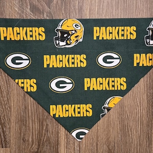 Könnte beinhalten: Grünes und gelbes Packers-Bandana. Das Bandana zeigt die Worte "PACKERS", das Logo des Teams und eine Footballhelm-Grafik. Das Bandana besteht aus einem weichen Stoff und ist dreieckig geformt.