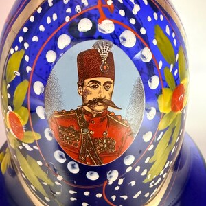 May include: A blue glass decorative bottle with a portrait of a man in a red uniform. The bottle has white polka dots, gold accents, and floral designs. The portrait is in a light blue circle.