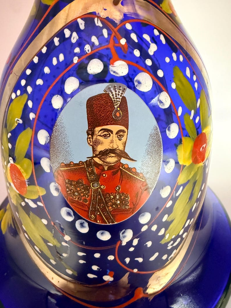 May include: A blue glass decorative bottle with a portrait of a man in a red uniform. The bottle has white polka dots, gold accents, and floral designs. The portrait is in a light blue circle.
