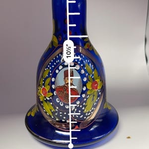 May include: A cobalt blue glass vase, approximately 10.5 inches tall, with a flared base and a narrow neck. The vase features a painted portrait and floral accents in gold, red, and yellow.