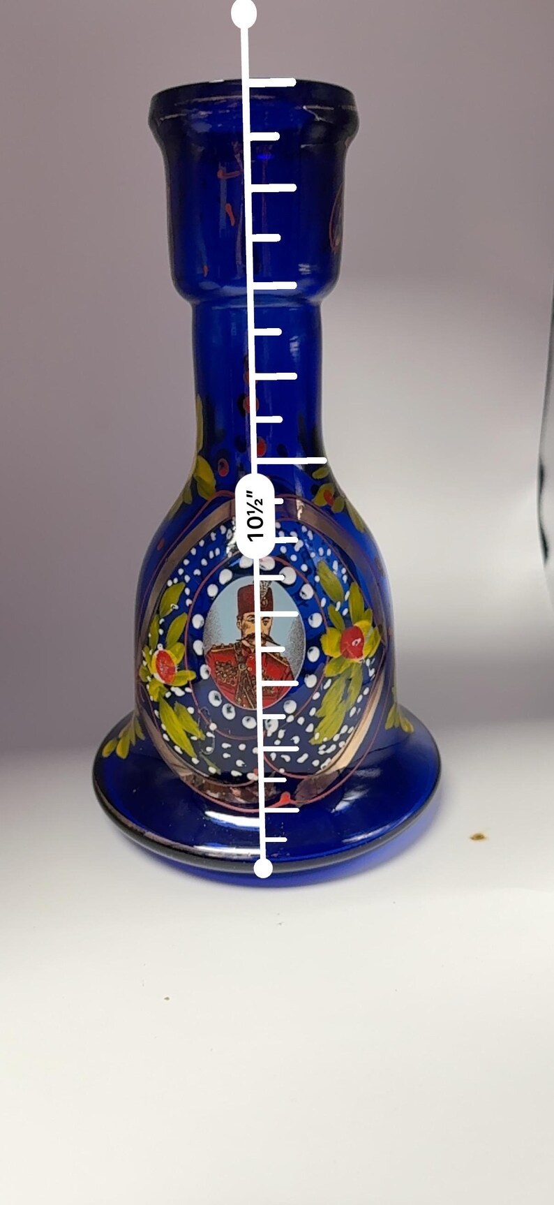 May include: A cobalt blue glass vase, approximately 10.5 inches tall, with a flared base and a narrow neck. The vase features a painted portrait and floral accents in gold, red, and yellow.