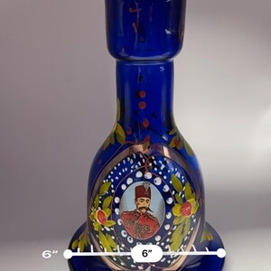 May include: A cobalt blue glass decorative bottle, approximately 6 inches in diameter, featuring a portrait of a man in a red coat and a decorative floral design. The bottle has a flared base and a cylindrical top.
