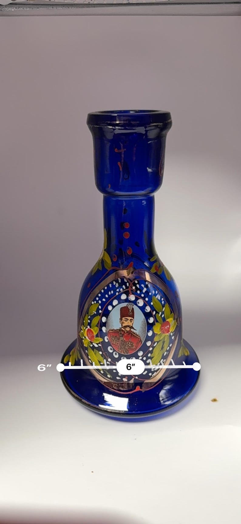 May include: A cobalt blue glass decorative bottle, approximately 6 inches in diameter, featuring a portrait of a man in a red coat and a decorative floral design. The bottle has a flared base and a cylindrical top.