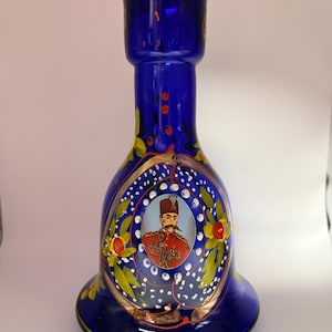 May include: A cobalt blue glass vase with a flared base and a bulbous body. The vase features a portrait of a man in a red uniform, surrounded by floral and gold accents. The vase is approximately 10 inches tall.