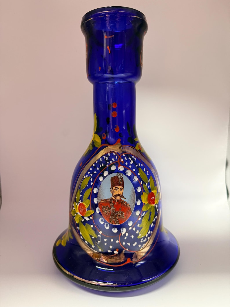 May include: A cobalt blue glass vase with a flared base and a bulbous body. The vase features a portrait of a man in a red uniform, surrounded by floral and gold accents. The vase is approximately 10 inches tall.