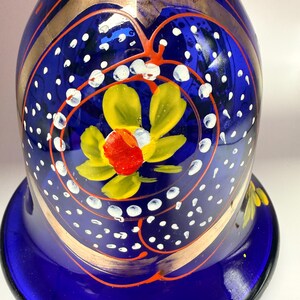 May include: A cobalt blue glass vase with gold accents and hand-painted floral designs. The vase features a flared base and a rounded body, with yellow and red flowers. White polka dots and orange swirls add detail.
