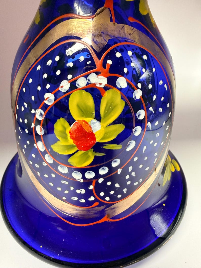 May include: A cobalt blue glass vase with gold accents and hand-painted floral designs. The vase features a flared base and a rounded body, with yellow and red flowers. White polka dots and orange swirls add detail.