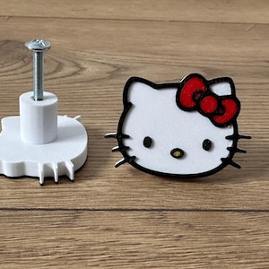 Może przedstawiać: Biały plastikowy gałka do szafki z wzorem Hello Kitty. Gałka ma czarny kontur i czerwoną kokardkę. Gałka jest przymocowana do srebrnej śruby.